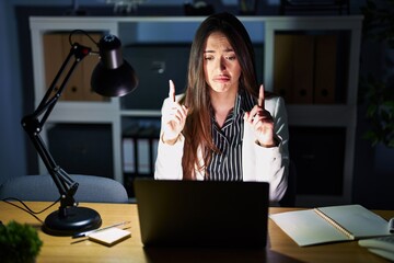 Poster - Young brunette woman working at the office at night with laptop pointing up looking sad and upset, indicating direction with fingers, unhappy and depressed.