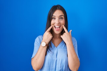 Poster - Young brunette woman standing over blue background smiling with open mouth, fingers pointing and forcing cheerful smile