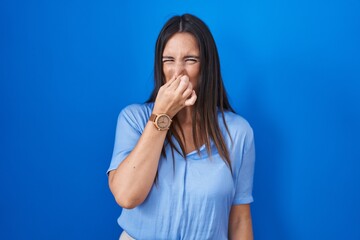 Poster - Young brunette woman standing over blue background smelling something stinky and disgusting, intolerable smell, holding breath with fingers on nose. bad smell