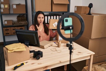 Wall Mural - Young latin woman working at small business ecommerce doing video covering mouth with hand, shocked and afraid for mistake. surprised expression