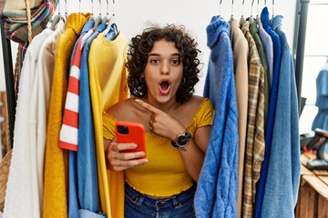 Sticker - Young hispanic woman searching clothes on clothing rack using smartphone surprised pointing with finger to the side, open mouth amazed expression.