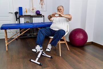 Sticker - Senior caucasian man at physiotherapy clinic using pedal exerciser smiling in love doing heart symbol shape with hands. romantic concept.