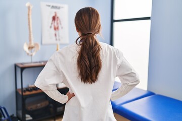 Wall Mural - Young brunette woman working at pain recovery clinic standing backwards looking away with arms on body