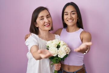 Wall Mural - Hispanic mother and daughter holding bouquet of white flowers smiling cheerful offering palm hand giving assistance and acceptance.