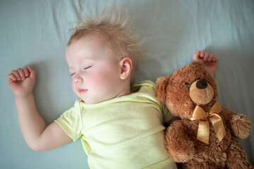 Wall Mural - small child sleeps in bed with teddy bear. dishevelled child with blond hair lies closing eyes in bed on blue sheet.