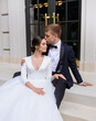 Portrait of tenderness married couple groom kissing forehead wife in beautiful white dress. Loving couple sitting on stairs enjoying the wedding day ceremony. Marriage. Family