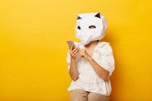 Portrait Of Anonymous Woman Blogger Wearing Dress And Paper Cat Mask Holding Cell Phone, Writing Post For Her Blog, Using Mobile Phone, Posing Isolated Over Yellow Background.