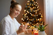 Smiling beautiful attractive young adult woman wearing white shirt sitting on table near christmas tree and holding mobile phone in hands, writing Xmas greeting in social networks.