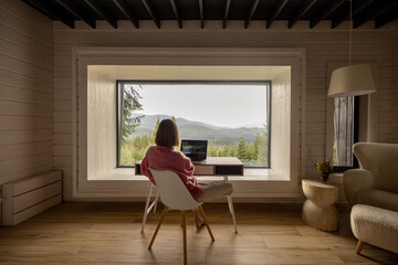 Woman works on laptop while sitting by the table in front of panoramic window with great view on mountains. Wide interior view. Remote work and escaping to nature concept