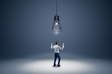 Back view of young businessman with abstract huge lamp hanging above. Idea, innovation and brainstorm concept.
