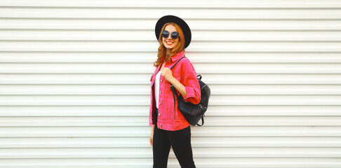 Portrait of stylish modern smiling young woman wearing pink jacket, black round hat and backpack on white background, blank copy space for advertising text