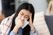 © kucherav - Young sick Asian woman sits on the sofa blowing her nose. Runny nose, cold or flu disease concept