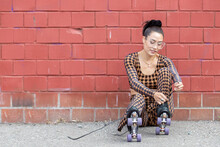 Asian Woman Wearing Checkered Pattern Body Suit Ties The Laces On Her Roller Skates While Sitting On The Ground Against A Red Brick Wall Outside.