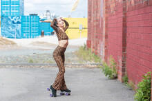 Asian Woman Wearing Roller Skates With Colorful Wheels Stands Near Brick Wall Outside With Her Body Outstretched In A Checkered Tight Fitting Outfit