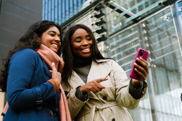 Two young cheerful women using cellphone and pointing while walking outdoors