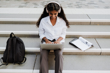 Wall Mural - Young woman using laptop and headphones while studying outdoors