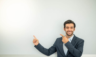 Portrait of a smiling young caucasian businessman wearing a dark suit and presenting copy space on his pointing finger near with concrete wall.