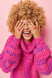 Overjoyed curly haired young European woman covers eyes with hand laughs happily smiles broadly hides her genuine emotions wears knitted jumper isolated over pink background. Happiness concept