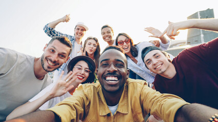 Point of view shot of handsome African American man taking selfie with his happy friends, people are looking at camera, smiling and posing with bottles at funny roof party.