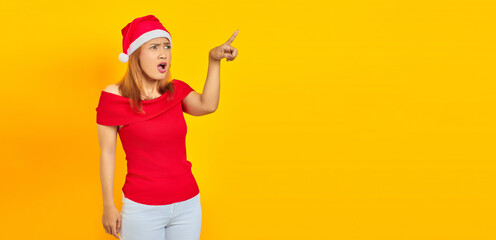 Wall Mural - Surprised young Asian woman wearing Christmas hat and pointing sideways over yellow background