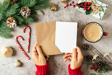Desktop Of Woman Hands In Red Sweater Holding Blank Paper And Envelope . Flat Lay Of Gray Background With Cup Of Coffee And Christmas Decoration. Top View Mock Up And Copy Space For Text.