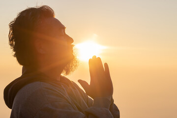 Close up portrait of young adult male with beard with praying hands praying for thank god golden sunset background. Concept of religion faith