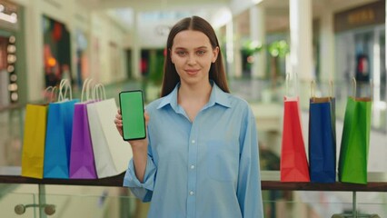 Smiling woman holding smartphone, showing green screen phone to camera in the shopping mall. Chroma key smartphone. Green screen technology. Sale and discount store