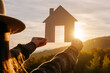 © Yura Yarema - Close up of young woman in hat holding paper house against sunset or sunrise light on mountains background. Home in rays of sun in autumn nature. Family life, mortgage, business real estate concept