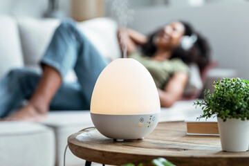 Essential oil aroma diffuser humidifier diffusing water articles in the air while woman listening music lying on coach.