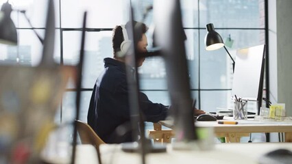 Fototapete - Portrait of Asian Creative Man Using Computer and Headphones in Bright Modern Office. Male Data Analyst Developing Database While Listening to Music. Active Dolly Shot from Behind a Desk.