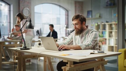 Fototapete - Portrait of Motivated Caucasian Young Man With Beard Working on a Laptop in Office. Male Director Preparing a Marketing Project. Diverse Team Working on Computers in the Background. Zoom In Shot.