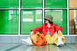 © okrasiuk - Hipster young woman in bright clothes, sun glasses, backpack bag and bucket hat posing while sitting near green glass wall background. Urban city street fashion. Fashion blogger. Selective focus.