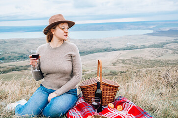 Young woman with glass of wine having picnic in mountains on autumn day, back view