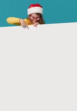 Cute Little Girl In A Santa Claus Hat And Party Glasses On A Turquoise Background. The Child Holds A Sign For Advertising, Empty Space For Text. Vertical Banner
