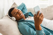 © Prostock-studio - Glad millennial african american guy lies on sofa in wireless headphones, typing on phone, watch video