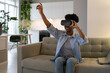 © DimaBerlin - Young smiling excited African American woman using VR glasses for online shopping in virtual reality, sitting on sofa in living room, black female exploring products virtually in augmented reality