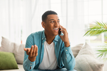 Wall Mural - Smiling millennial african american male gesturing hand and calling by smartphone in white living room interior