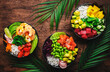 Hawaiian poke bowl set: tuna, salmon, shrimp with avocado, mango, radish, rice and other ingredients. Soy sauce and sesame dressing. Wooden table background, palm leaves, top view