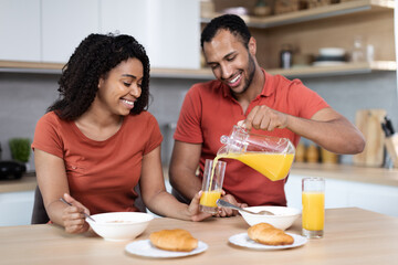 Wall Mural - Happy handsome young black guy in red t-shirt pours juice into glass to his wife, couple enjoys breakfast