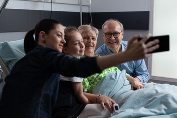 Wall Mural - Adult woman taking self portrait with smartphone accompanied of elderly patient, relatives in geriatric hospital room. Family selfie with sick grandmother at clinic visiting area.