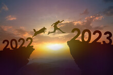 Silhouette Dog And Young Man Jumping From 2022 To 2023 At Cliff On Sunset. New Year And Success Concept.