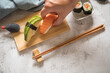 Frau isst Lachs Avocado Sushi als Nigiri, Maki, roh auf Holz Teller mit Soja Soße in Fisch Verpackung und Stäbchen auf Stein Hintergrund hell