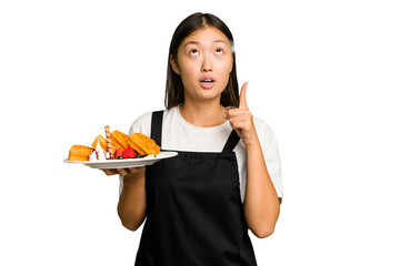 Wall Mural - Young asian waitress woman holding waffles isolated pointing upside with opened mouth.