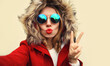 © guas - Close up portrait of stylish woman blowing her lips sends air kiss stretching hand for taking selfie with smartphone wearing red jacket with fur hood