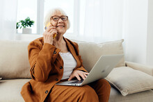 A Joyful Elderly Woman, A Businesswoman Works Remotely While In A Comfortable Environment At Home Holding A Laptop On Her Lap Solving Work Issues With A Pleasant Smile On Her Face