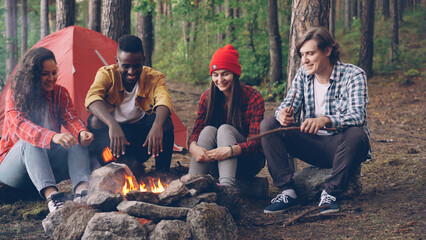 Multiethnic group of friends tourists are sitting around fire talking and laughing, young man is throwing firewood in flame. Camping, friendship and summer nature concept.