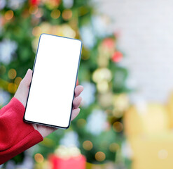Woman hand wearing red sweater holding a smartphone with empty isolated white display screen, with blurred decorating Christmas tree in bright cozy room.