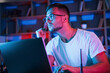 Man in glasses and white shirt is sitting by the laptop in dark room with neon lighting