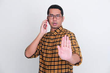 Wall Mural - Adult Asian man give stop hand sign with serious expression while answering a phone call