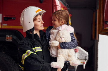 Kid is holding bunny toy. Firefighter woman in uniform is with a little girl